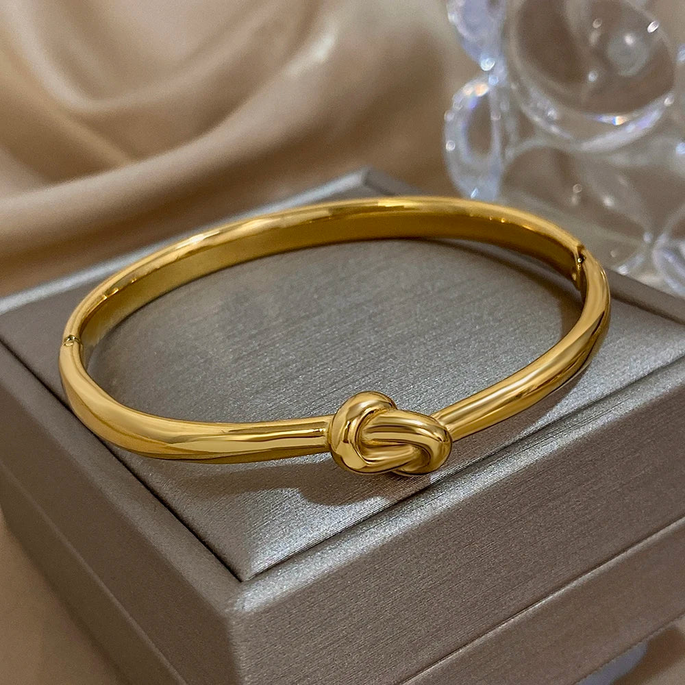 Simple thin stainless steel knotted bangle in gold color displayed on soft satin fabric.