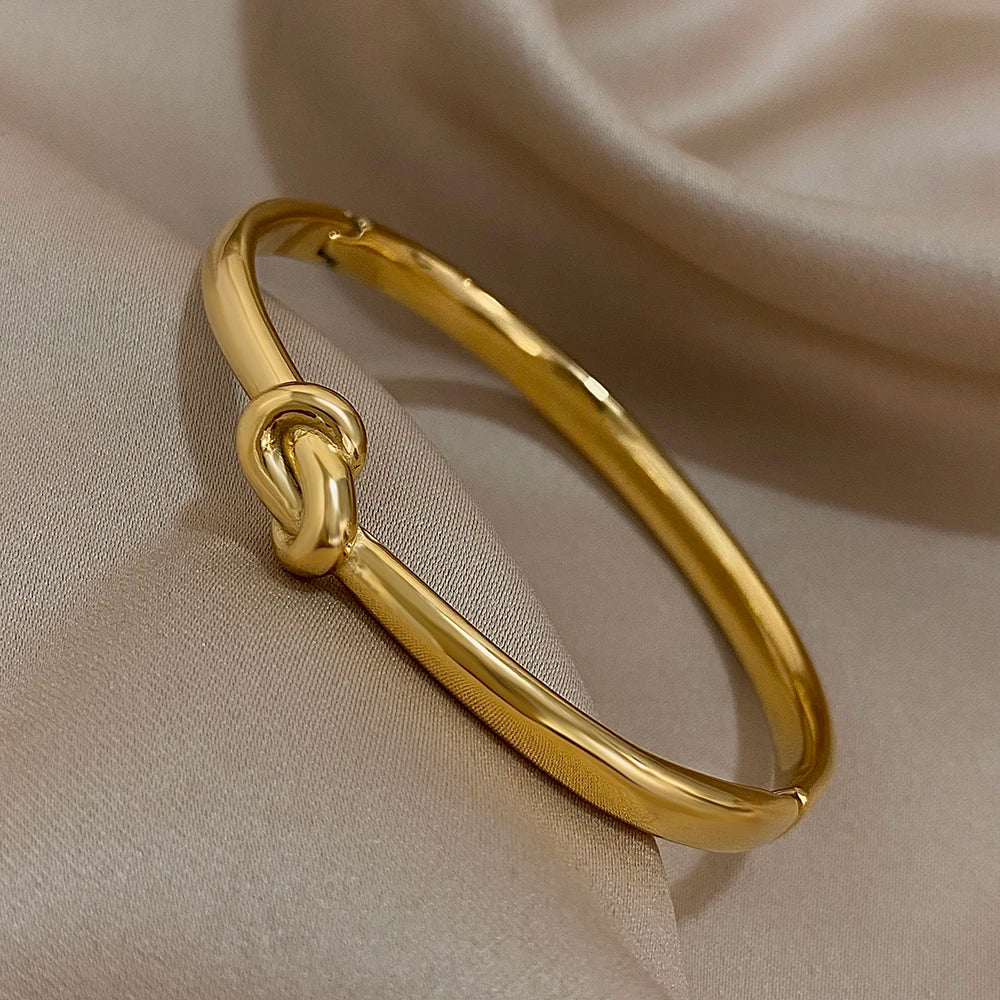 Simple thin stainless steel knotted bangle in gold color displayed on soft satin fabric.