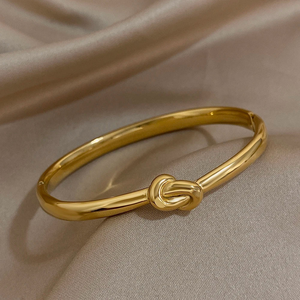 Simple thin stainless steel knotted bangle in gold color displayed on soft satin fabric.